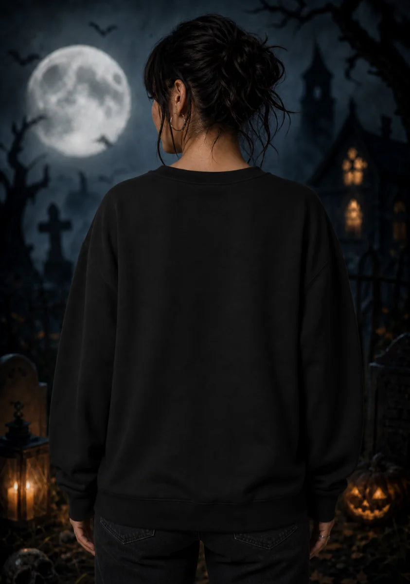 A woman stands with her back to the camera wearing a plain black sweatshirt in a moonlit graveyard with lanterns and pumpkins.