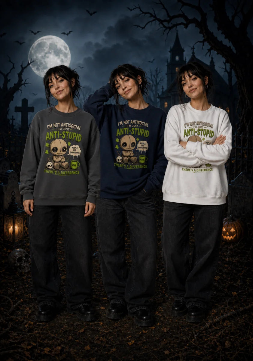 A woman stands in a spooky graveyard under a full moon wearing three colour variants of the “Anti‑Stupid” chibi sweatshirt.