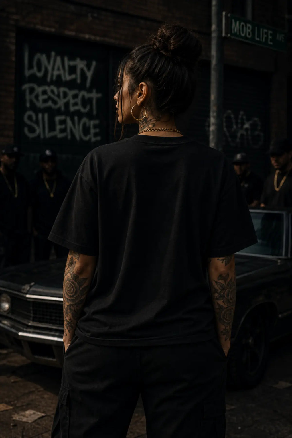 A person seen from behind in a black oversized T‑shirt and cargo pants standing on a city street with a car and graffiti behind them.