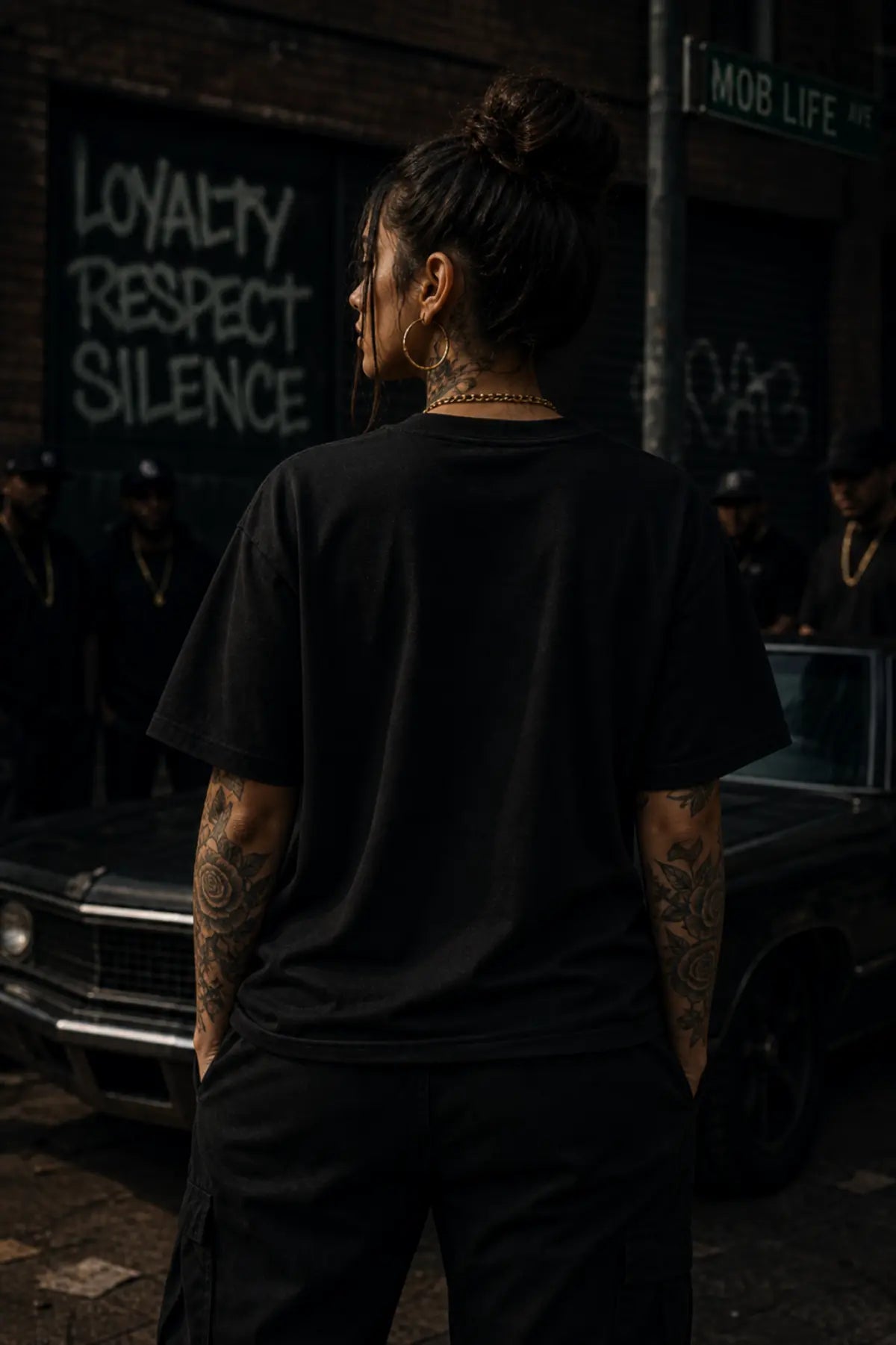 A person seen from behind in a black oversized T‑shirt and cargo pants standing on a city street with a car and graffiti behind them.