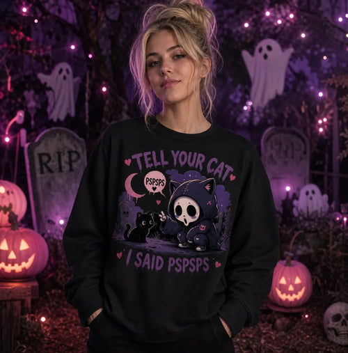 Person in Halloween graveyard scene wearing black sweatshirt with chibi skeleton‑cat design, amid pumpkins, ghosts and purple glow