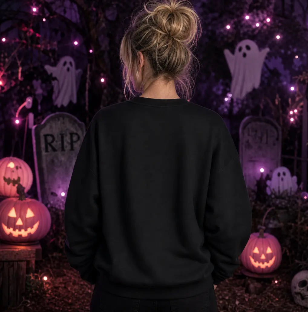 Person facing away in Halloween scene wearing black sweatshirt, standing amid jack‑o’‑lanterns, ghosts, tombstones and purple lights