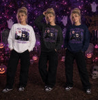 Three people in Halloween scene wearing chibi skull‑cat sweatshirts, standing amid tombstones, pumpkins, ghosts and purple lights