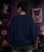 Person in dark blue sweatshirt and denim shorts facing away, surrounded by gothic plush toys, candles, and eerie purple lighting.