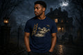 Man in a navy blue graphic t‑shirt with a ghost and ladder design standing in a foggy graveyard at night near a haunted house
