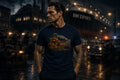 A man in a navy graphic T‑shirt stands in a rainy dark industrial port with an RWI Exports ship visible in the background.