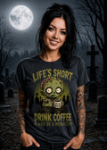 A woman stands in a moonlit graveyard wearing a black T‑shirt with a zombie graphic and “Life’s Short Drink Coffee” text.