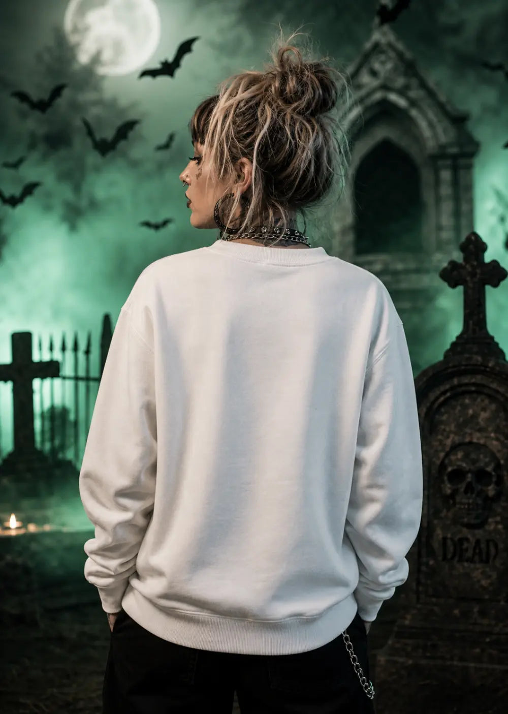 A female stands in a green‑lit graveyard wearing a plain white sweatshirt, surrounded by tombstones and eerie moonlight.