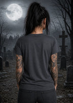 A woman stands in a moonlit graveyard wearing a dark T‑shirt, her tattooed arms visible against the eerie night backdrop.
