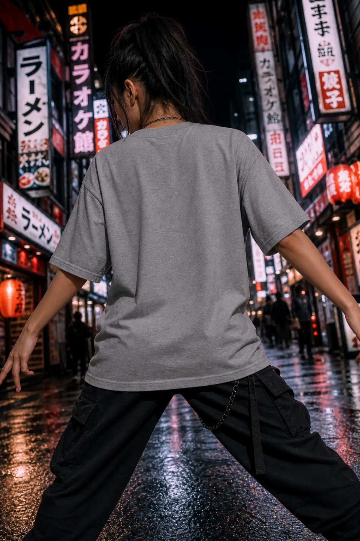 A person seen from behind in a gray short‑sleeve T‑shirt and black cargo pants on a wet night street with colorful neon signs.