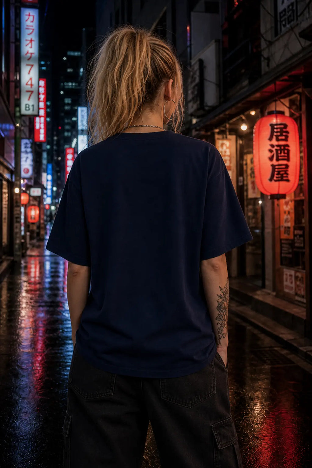 A person seen from behind wearing a dark navy short‑sleeve T‑shirt and black cargo pants on a wet night street with neon Japanese signs.